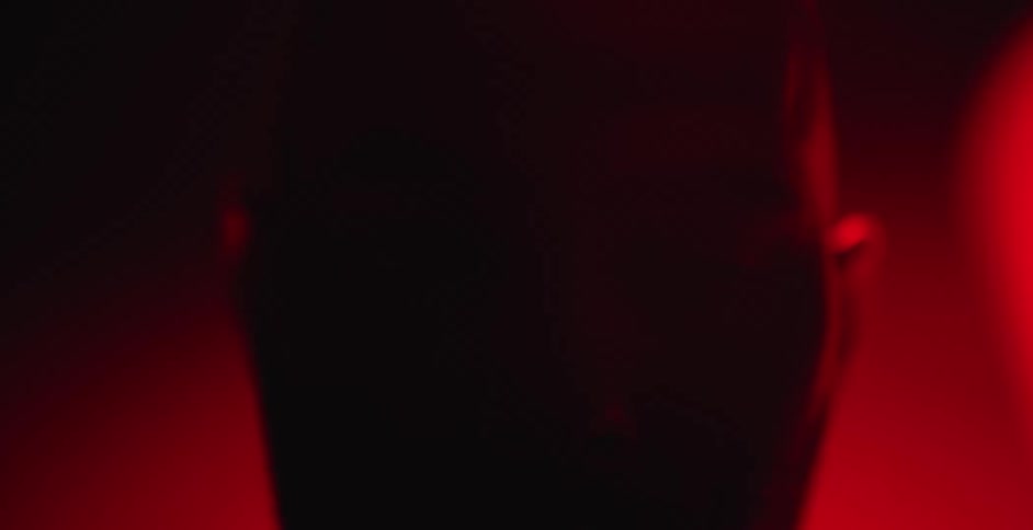A mysterious man's face is illuminated by a dramatic red light before being consumed by darkness