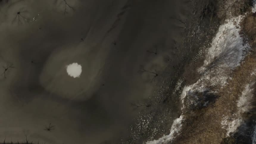 A slow, rotating top-down aerial view of a frozen lake reveals abstract patterns and cracks in the dark ice next to a snow-dusted shoreline
