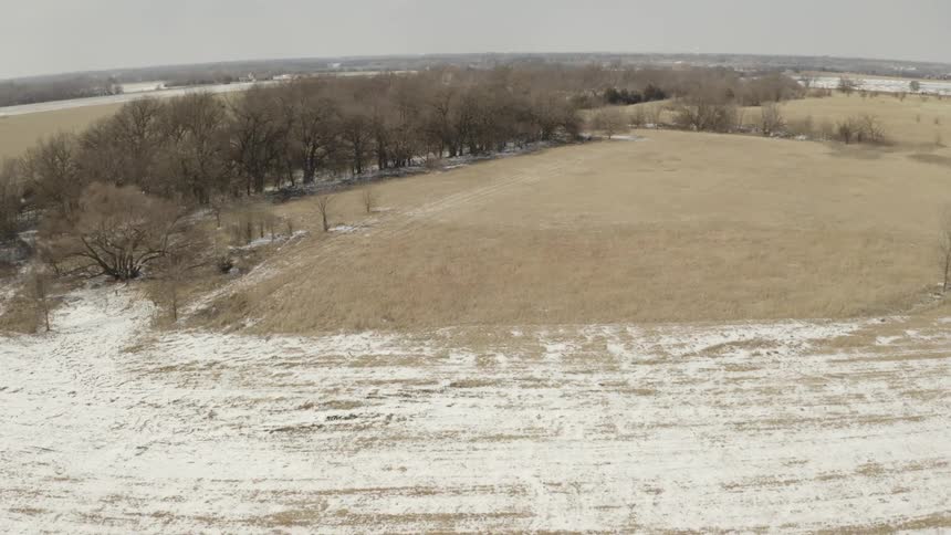 Aerial footage slowly advances over a snow-dusted prairie landscape with leafless trees under an overcast winter sky