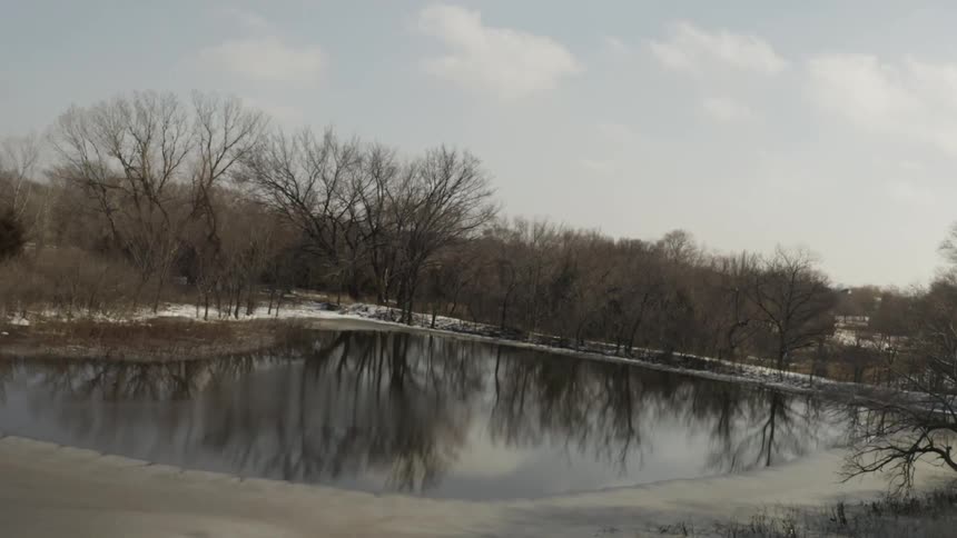 A slow, Dutch angle zoom over a partially frozen winter pond reflecting bare trees