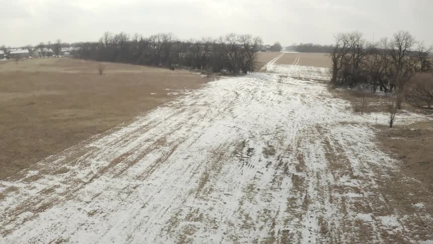 A slow aerial pan reveals a desolate winter landscape with snow-dusted brown fields and a line of bare trees on an overcast day