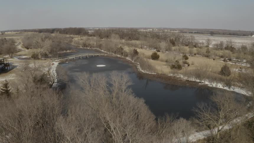 A slow aerial push-in over a partially frozen lake with a small bridge in a rural winter landscape