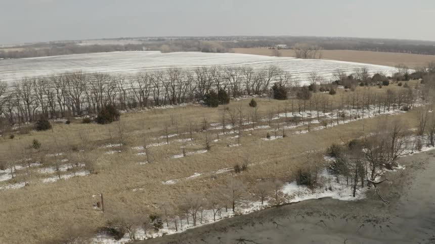 A slow aerial shot moves across a rural winter landscape featuring snow-dusted fields, bare trees, and a frozen pond in the foreground