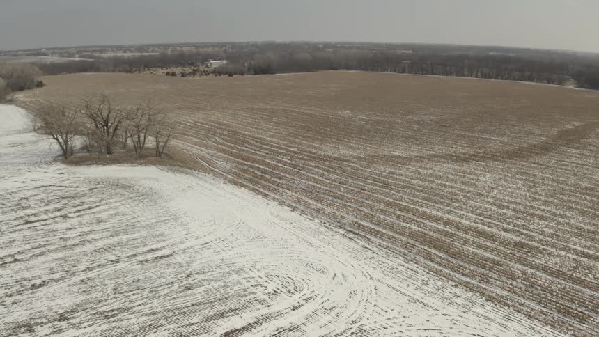 A slow aerial drone flight over a vast, snow-covered farm field on a bleak winter day