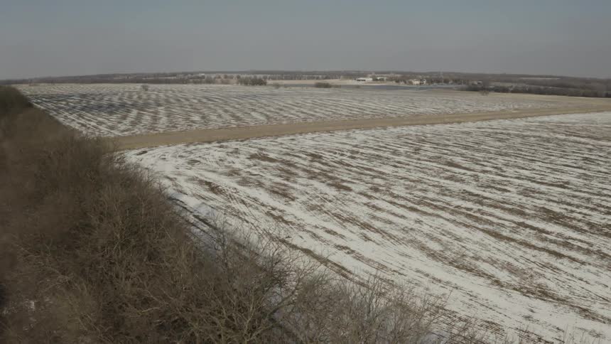 An aerial view reveals a vast, snow-dusted agricultural field behind a line of bare winter trees in the countryside