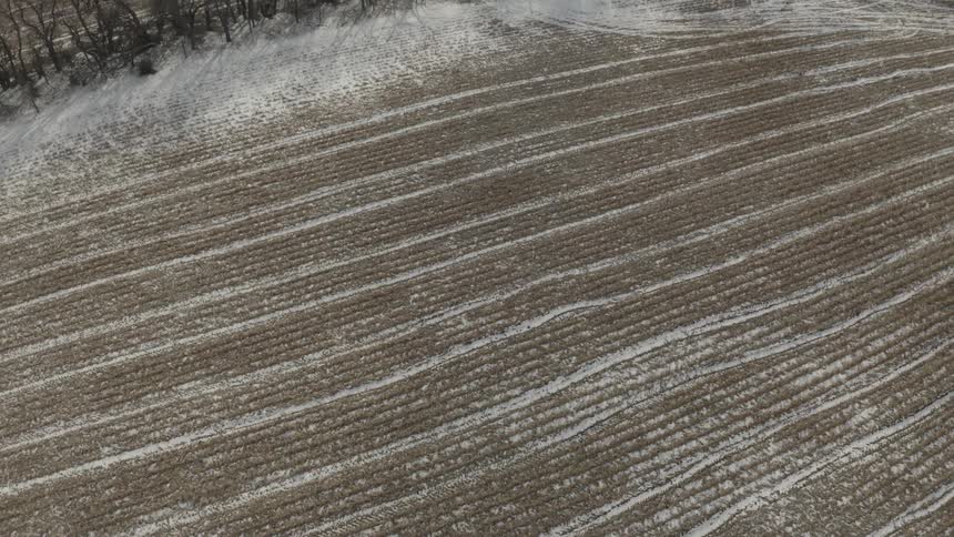 An aerial view pulls back from the snowy rows of a harvested farm field to reveal a stark winter landscape with bare trees