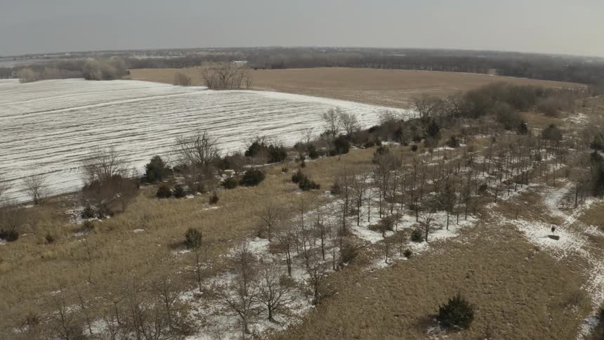 An aerial view slowly advances over a frozen pond with a bridge towards snow-dusted fields in a tranquil rural winter landscape