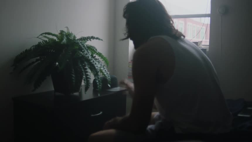 A man gestures his hands while listening to music on the edge of a bed