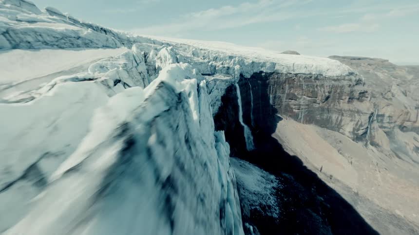 A high-speed FPV drone navigates the jagged ice spine of a vast glacier, revealing meltwater waterfalls cascading down the mountain
