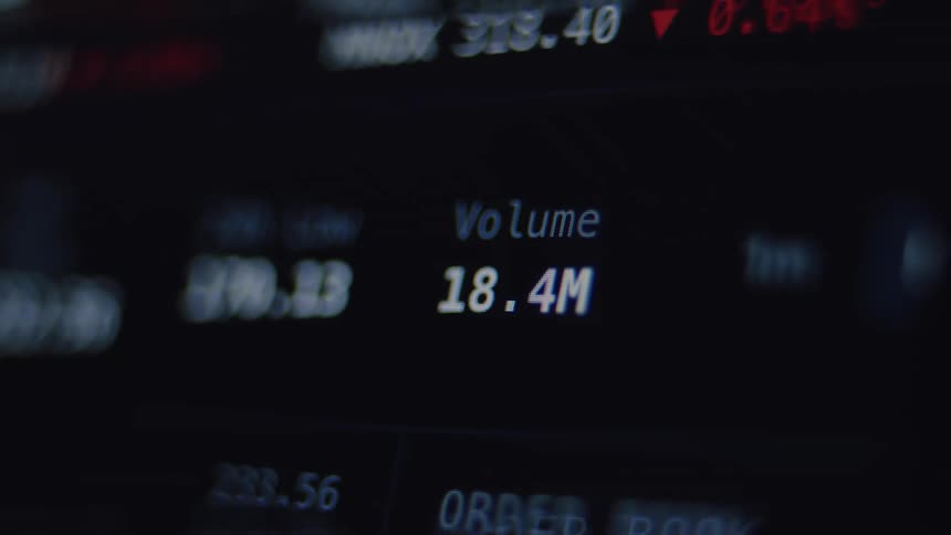 A close-up, out-of-focus shot of a digital stock market ticker screen shows fluctuating prices and trading volume