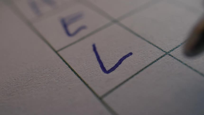 A macro close-up shot shows a ballpoint pen making a checkmark in a box on a gridded sheet of paper