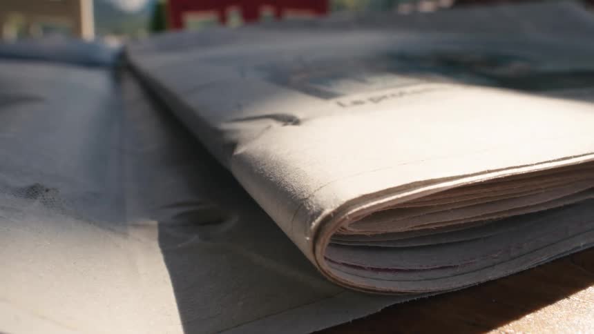 A close-up shot in slow motion shows a newspaper page being turned on a wooden table as sunlight moves across it