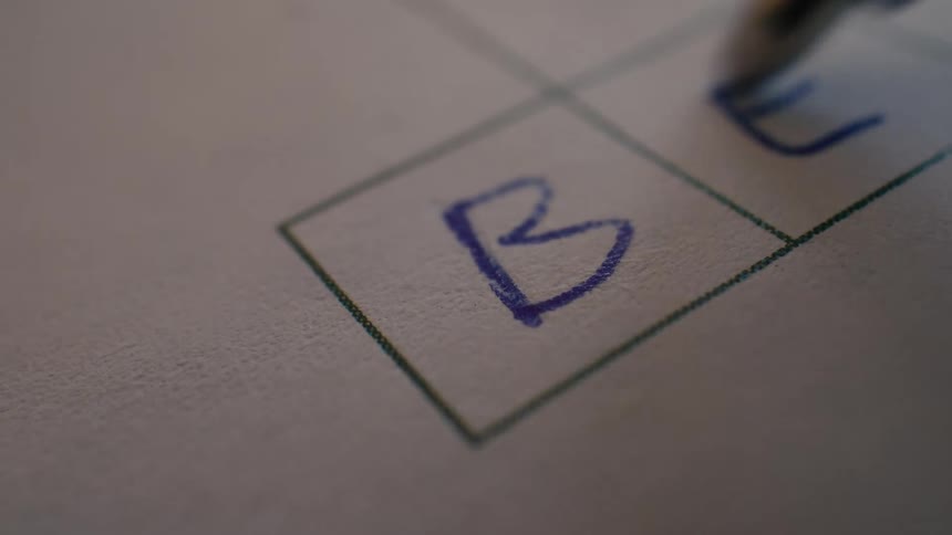 Extreme close-up shot of a blue ink pen writing the letters 'B' and 'E' into boxes on paper