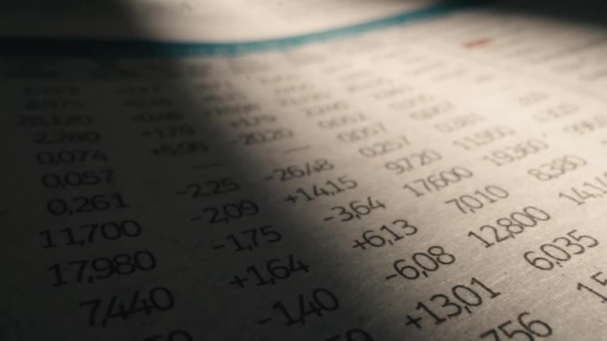 A dramatic close-up of stock market data printed in a newspaper, with light and shadow moving across the page