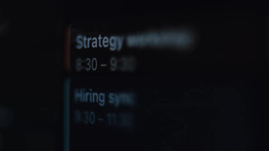 A macro close-up shot of a digital calendar on a screen showing a busy schedule being scrolled through in a dark setting