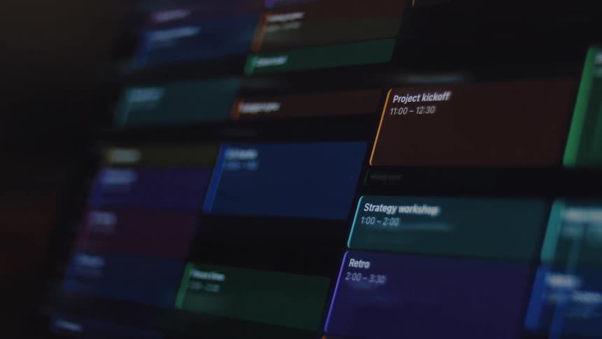 A close-up view of a digital project management calendar in dark mode being populated with color-coded events and meetings