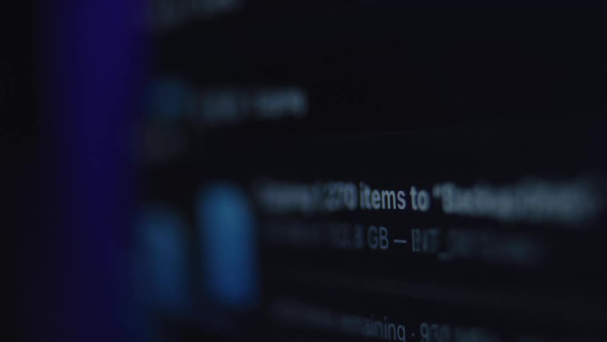 A close-up, out-of-focus shot shows a computer screen displaying a fast-paced file copying and data transfer process in a dark room