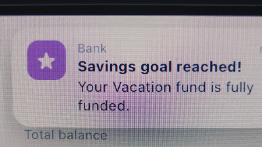 A mobile banking app notification on a digital screen shows that a vacation savings goal has been reached