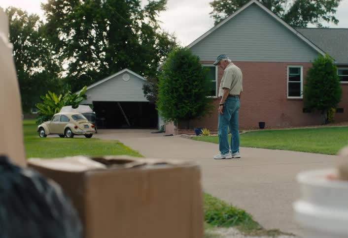A nostalgic older man stands in his driveway with moving boxes and a bucket of baseballs in the foreground