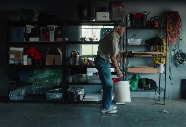 An elderly man gathers tools and supplies from a cluttered shelf in his garage, places them in a bucket, and walks away