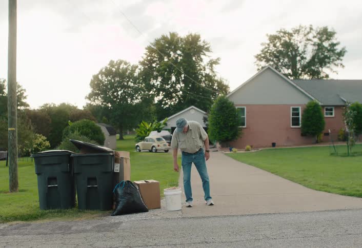An elderly man carries a bucket down his driveway to the curb for trash pickup before walking back towards his house in a quiet suburban neighborhood