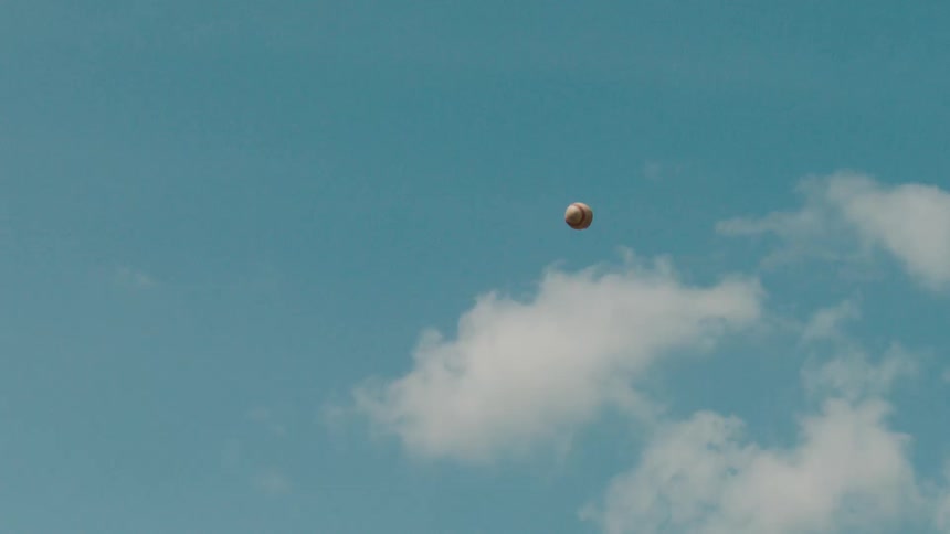 A slow-motion shot of a baseball tossed high into the air, flying and falling against a clear blue sky with white clouds