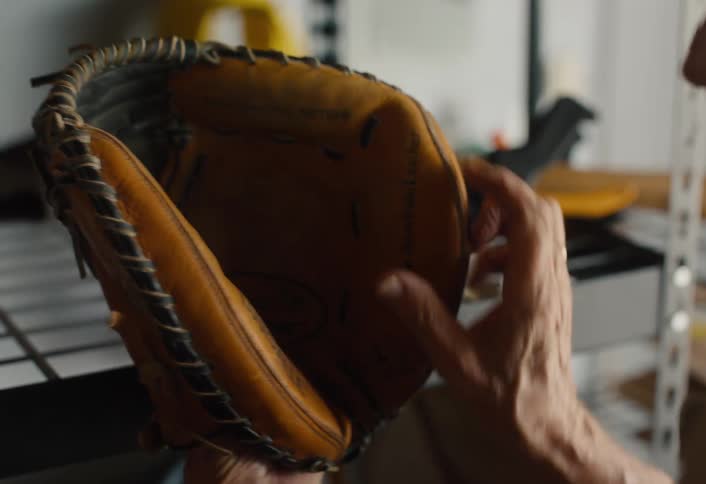 An older man rediscovers a well-worn baseball glove in a dusty garage and fondly punches his fist into it, reminiscing about the past