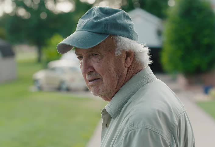 A sad elderly man wearing a baseball cap turns to look back with a thoughtful expression while standing outside his home
