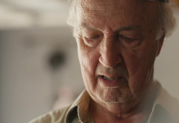 A close-up reveals the thoughtful face of an elderly man as he removes his cap in a softly lit workshop