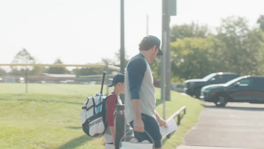 A father and son walk and talk along a path by a baseball field, heading home after practice on a sunny day