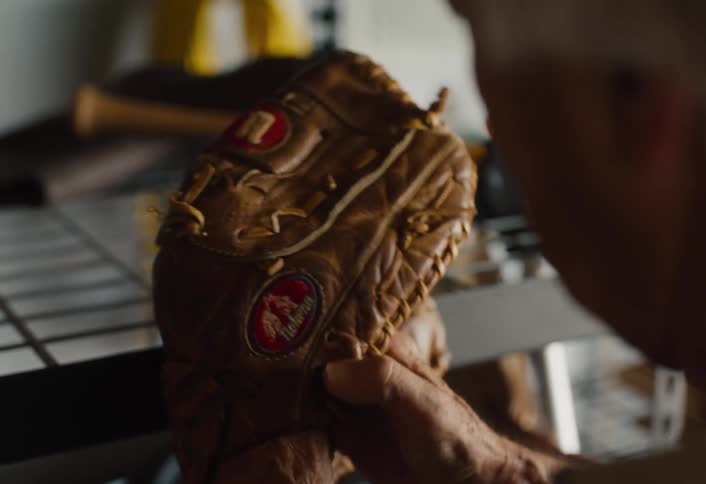 An older man fondly inspects his well-used leather baseball glove, breaking it in with his hands in a dimly lit storage space