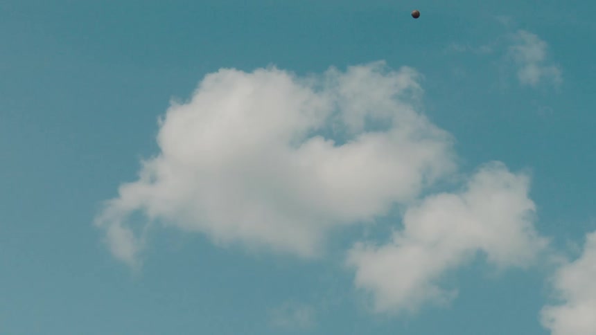 A baseball flies up and down through the air in slow motion against a blue sky with clouds