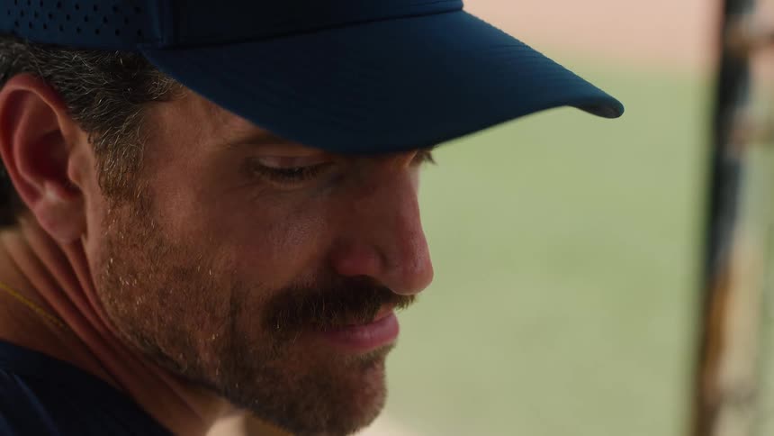 A close-up profile of a mustached baseball coach in a cap smiling thoughtfully from the dugout