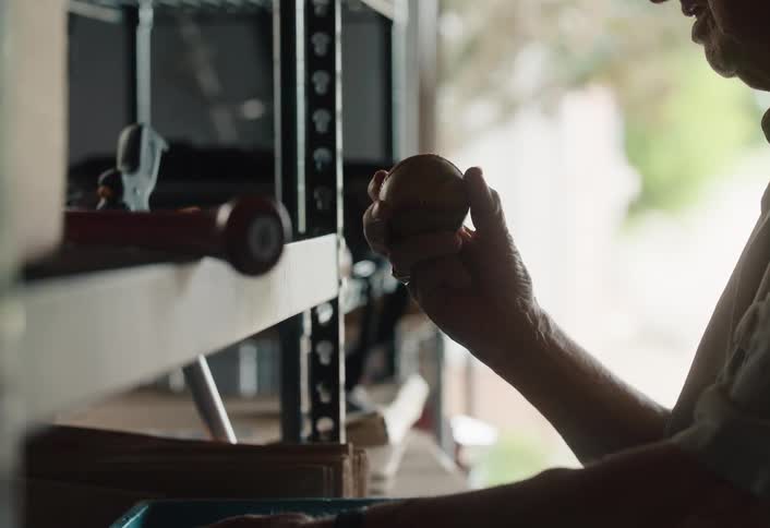 An elderly person's hands hold an old baseball, reflecting on memories in a dimly lit garage