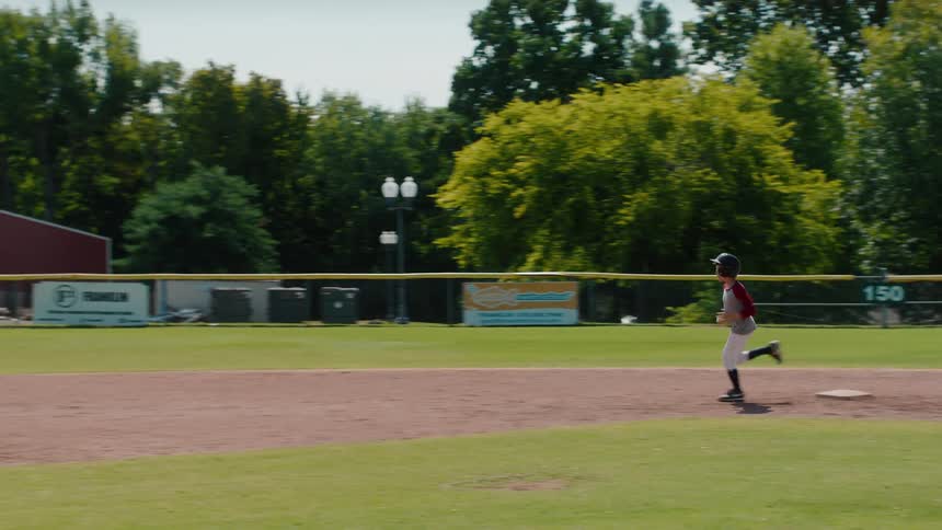 A young boy in a baseball uniform runs the bases in slow motion on a sunny day