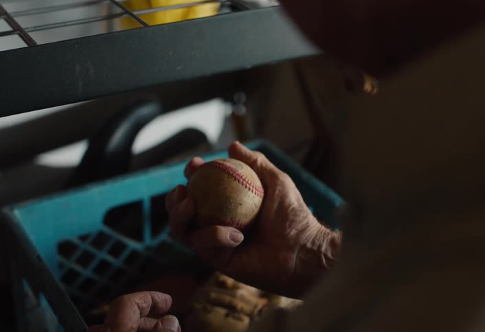 The weathered hands of an elderly man find an old, worn baseball in a dusty crate, holding it while reminiscing about past memories