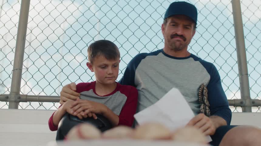 A baseball coach sits on the bench with his disappointed son, offering encouragement before they grab a bucket of balls to go practice