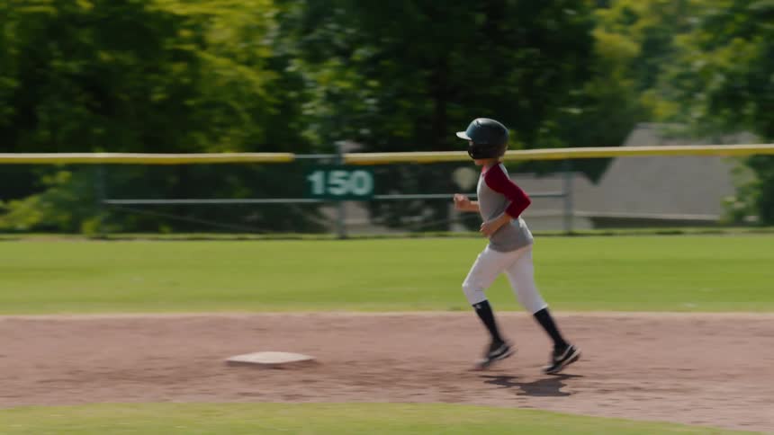 A determined young boy in a helmet and uniform runs the bases in slow motion during a Little League baseball game on a sunny day