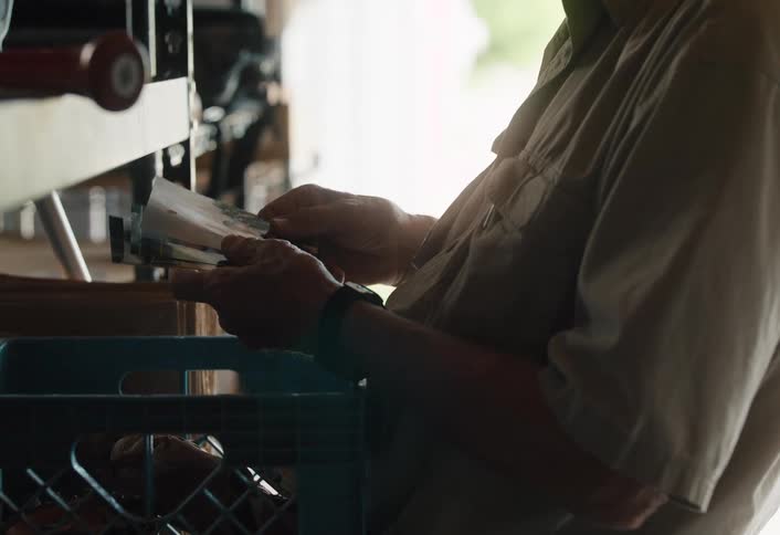 An older man stands in his garage, backlit by natural light, as he nostalgically looks through a stack of old printed photographs