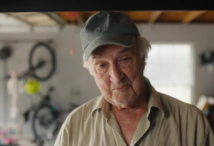 An elderly man in a cap organizes his garage, lifting and placing a blue crate on a high shelf in slow motion