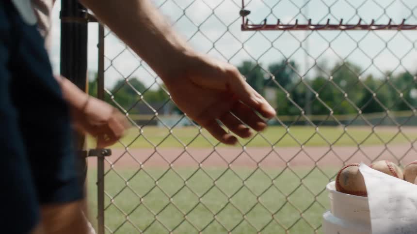 A father and son walk into the dugout on a baseball field where the father reads a note on a bucket of baseballs