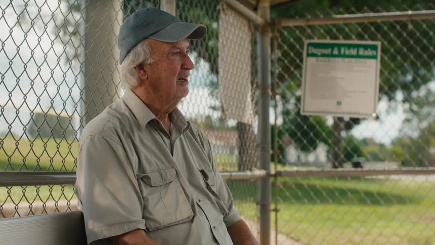 An elderly man sits alone on a bench in a baseball dugout, watching a game with mixed emotions