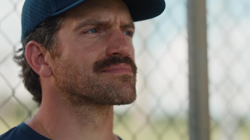 Close up on a tired but hopeful baseball coach leaning against a chain-link fence