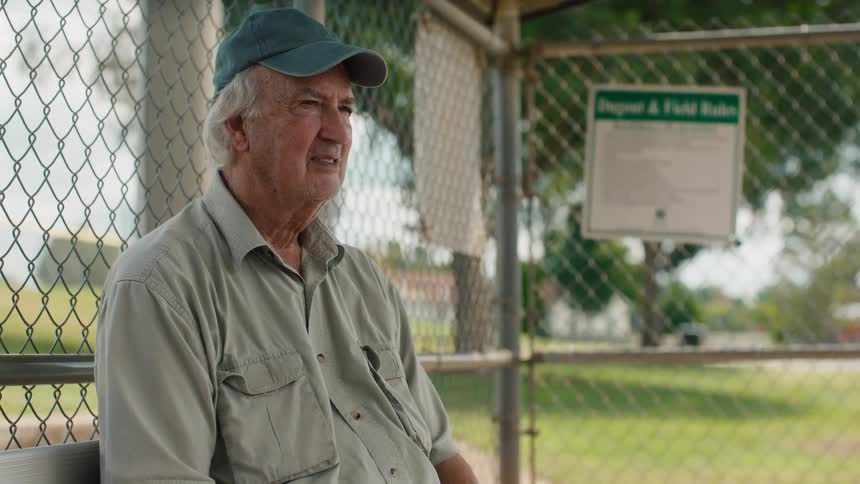 A nostalgic older man sits alone on a dugout bench, thoughtfully watching a baseball game at a local park