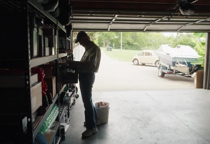 A man organizes items on his garage shelves before picking up a bucket and walking outside into the daylight