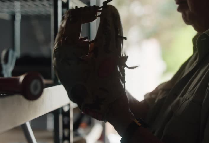 An older man fondly remembers his past as he holds his old, well-worn baseball glove in a dimly lit garage