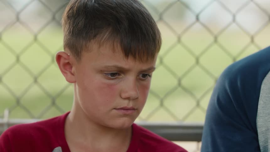 A close-up of a dejected young boy sitting in a dugout, looking down with a sad expression before glancing up