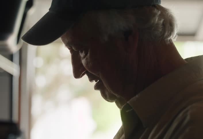 An elderly man in a cap smiles nostalgically while looking through a stack of old family photographs in a softly lit garage