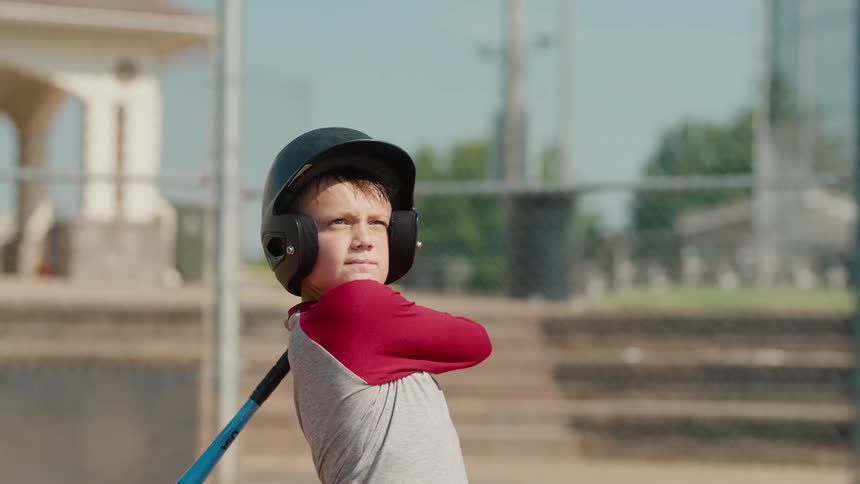A focused young boy in a batting helmet swings a baseball bat in slow motion and begins to run on a sunny day