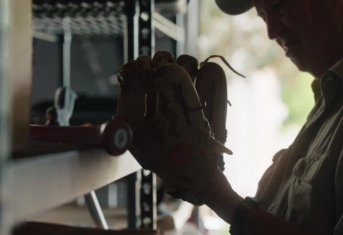 An older man fondly remembers past games while carefully inspecting his well-worn baseball glove in a dimly lit garage